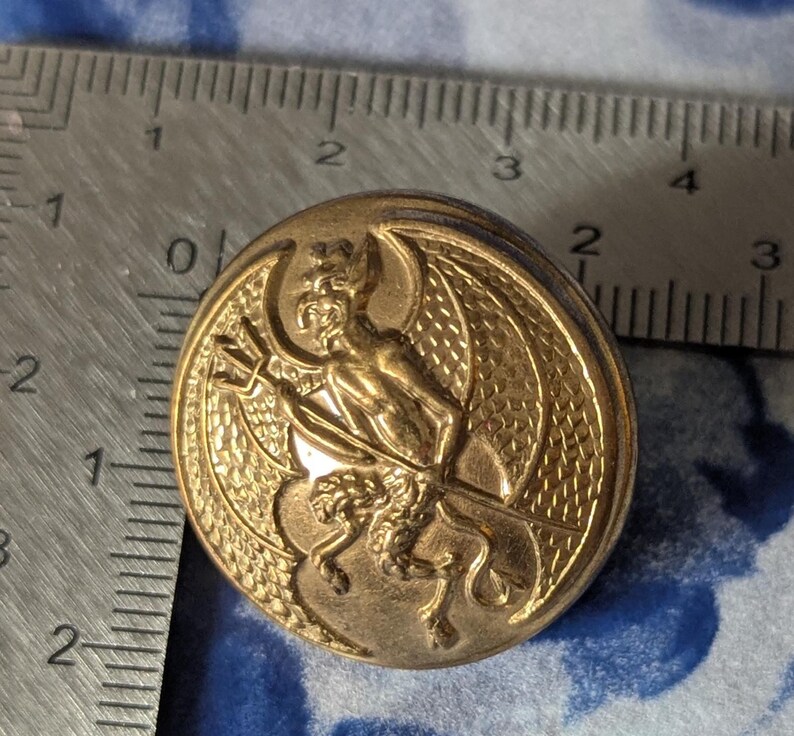 May include: A gold-colored button with a detailed embossed design of a devil figure holding a trident. The devil has wings and is seated on a horned creature. The button is round and has a diameter of approximately 1 inch.