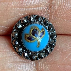Op de afbeelding: Een ronde knoop met een felblauw middenstuk en een geschilderd paars en goudkleurig bloemmotief. Het centrale ontwerp is omringd door een rand van kleine, donkere, gefacetteerde metalen kraaltjes die in een ring langs de rand liggen.