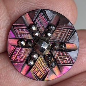 以下が含まれることがあります： 中央にファセットカットの宝石と幾何学模様が施された、丸い虹色のボタン。ボタンは、紫、茶色、金色の濃いマルチカラーの表面が特徴です。小さなファセットカットのビーズが中央の宝石の周りに配置されています。