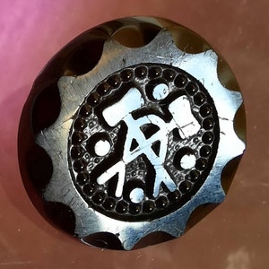 Puede incluir: Un botón marrón oscuro con un diseño único. El botón tiene un borde festoneado y un diseño central con un símbolo blanco rodeado por un círculo de cuentas. La superficie del botón es brillante.