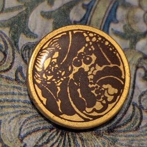 Op de afbeelding: Een ronde knoop in antieke stijl met een goudkleurige rand en een donkerbruin midden. Het midden heeft een gedetailleerd bloemenontwerp in een lichtere goudkleur. De knoop is geplaatst tegen een patroonachtergrond.