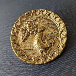 Puede incluir: Un botón de metal dorado con un diseño en relieve de un gallo con un racimo de uvas. El botón tiene un borde decorativo con pequeños círculos de espejo.