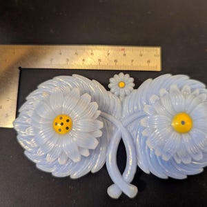 Puede incluir: Broche en forma de margarita azul claro con centro amarillo y puntos negros. El broche mide aproximadamente 10 cm de ancho. Tiene un diseño detallado de pétalos y está hecho de un material translúcido.