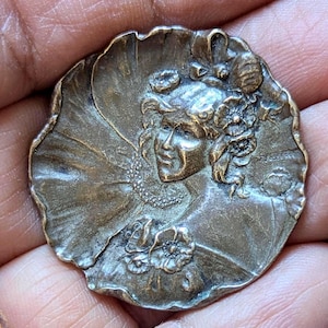 May include: A circular bronze-toned metal brooch featuring a relief sculpture of a person with wavy hair adorned with flowers. The design includes a ruffled collar, floral accents at the base, and the engraved text F. LASSERRE on the right side.