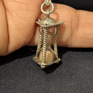 Op de afbeelding: Een zilverkleurige ketting met een zandloperhanger. De zandloper is gevuld met bruin zand en ingekapseld in een decoratief frame. De ketting is ook zilverkleurig. De hanger hangt aan een fijne ketting.