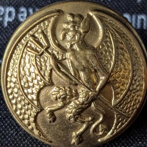 May include: A gold-colored button with a detailed embossed design. The central figure is a winged demon holding a trident, with a lion-like creature at its feet. The button has a textured, circular border.