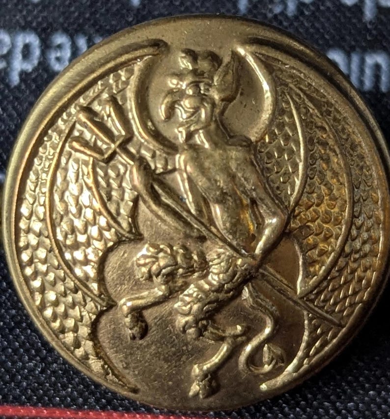 May include: A gold-colored button with a detailed embossed design. The central figure is a winged demon holding a trident, with a lion-like creature at its feet. The button has a textured, circular border.