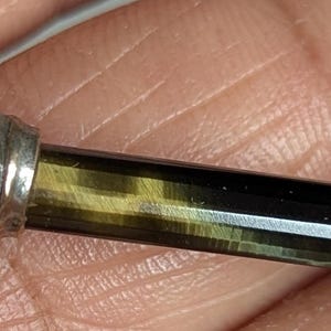 Peut inclure: Gros plan sur un stylo décoratif avec une extrémité en métal doré et un corps rayé noir et or. L'extrémité du stylo présente un motif orné. Le stylo est tenu dans une main.