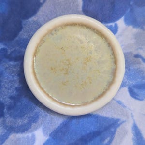 May include: A small, round ceramic dish with a cream-coloured rim and a pale yellow-green interior. The interior has a speckled, textured appearance. The dish is set against a blue and white floral patterned background.