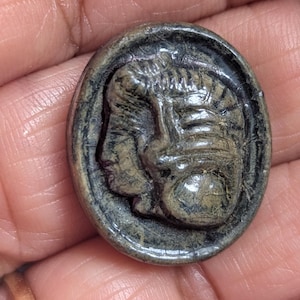 Pode incluir: Botão ou pingente oval escuro com um relevo detalhado de um perfil clássico. A figura tem um penteado e traços faciais detalhados. O botão é de um tom abafado e terroso, com textura e desgaste visíveis.