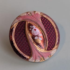 Peut inclure: Un bouton rond antique avec un centre tissé bordeaux et un motif floral rose et or. Le bouton a une bordure dorée et un fond texturé et tissé. Le motif floral présente des accents blancs et dorés.