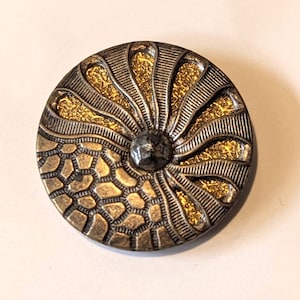 May include: A round, antique-style button with a detailed design. The button features a central, dark gray, faceted gem surrounded by a gold and bronze pattern. The outer edge has a textured, geometric design.
