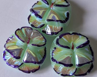 May include: Three glass beads shaped like pansy flowers. Each bead features a pale green base with iridescent highlights and deep purple painted edges on the petals. The centre of each flower has a small, raised, dark red detail.