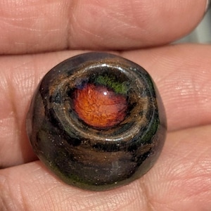 May include: A small, dark, rounded object with a glossy finish. The center features a vibrant orange and red design, surrounded by green and brown hues. The outer edges are dark, creating a striking contrast.