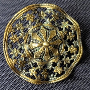 May include: An antique brass brooch with a floral design. The circular brooch features a central medallion with an intricate pattern, surrounded by a delicate openwork border of flowers and leaves. The brooch has a vintage aesthetic and is made of brass.