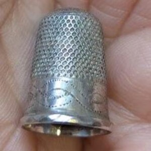 Op de afbeelding: Een zilverkleurige metalen vingerhoed met een gestructureerd honingraatpatroon op de bovenste koepel en een decoratieve band met een gegraveerd golvend lijnontwerp rond de basis. De vingerhoed heeft een gladde, gepolijste rand aan de onderkant.
