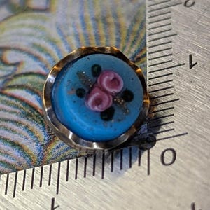 May include: A small, round button with a light blue center and a gold-colored metal rim. The center features pink floral designs and dark green accents. The button is approximately 0.5 inches in diameter.