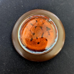 Könnte beinhalten: Ein runder, brauner Keramikuntersetzer mit einem glänzenden, gesprenkelten orangefarbenen Zentrum. Die Mitte hat dunkelbraune Flecken und ist von einem dünnen blau-weißen Ring umgeben. Der Untersetzer hat einen erhöhten Rand.