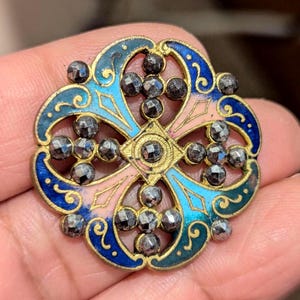 May include: A detailed, antique brooch with a cross design. The brooch features a gold-toned metal base with a central cross shape, adorned with small, dark gray stones. The design incorporates blue, pink, and green enamel, creating a colorful and intricate pattern. The brooch is a vintage piece.