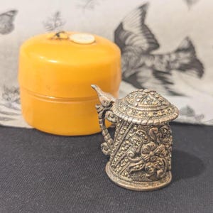 Puede incluir: Un dedal de plata con forma de jarra de cerveza con intrincados diseños florales y un asa en forma de pájaro. El dedal está sobre una superficie negra junto a un recipiente amarillo.