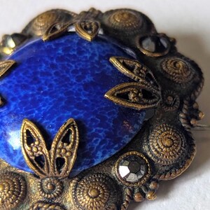 May include: Vintage brooch with a large, oval, speckled blue stone center. The stone is surrounded by an ornate, antique gold-toned metal frame with decorative filigree and small, faceted black stones. The brooch has a pin on the back.