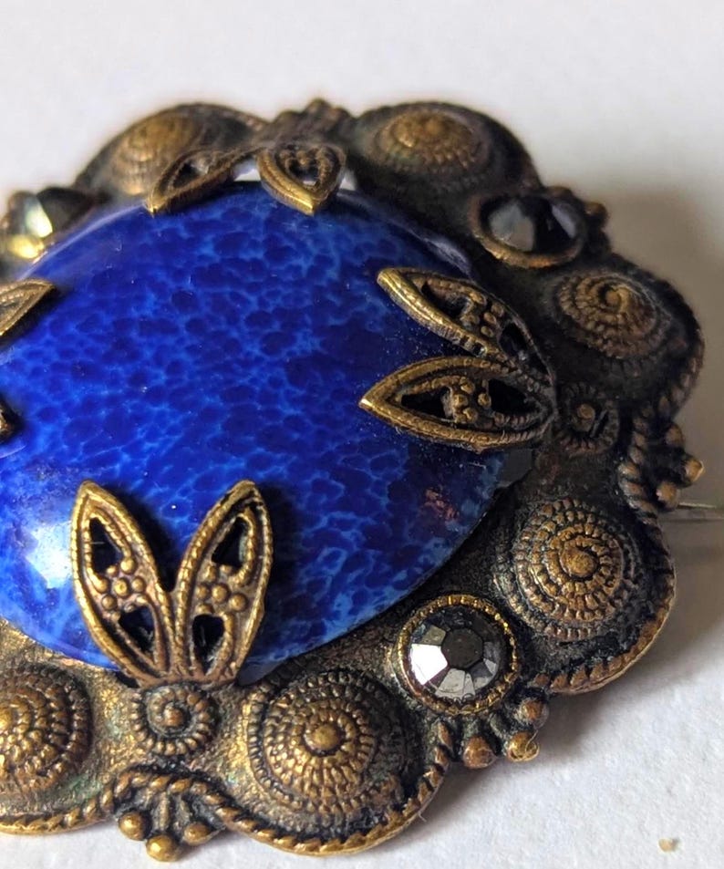 May include: Vintage brooch with a large, oval, speckled blue stone center. The stone is surrounded by an ornate, antique gold-toned metal frame with decorative filigree and small, faceted black stones. The brooch has a pin on the back.