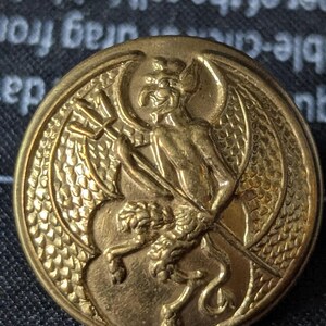 May include: A gold-colored button with a detailed relief of a devil-like figure holding a trident. The button has a circular shape with intricate detailing around the edges and the central image.