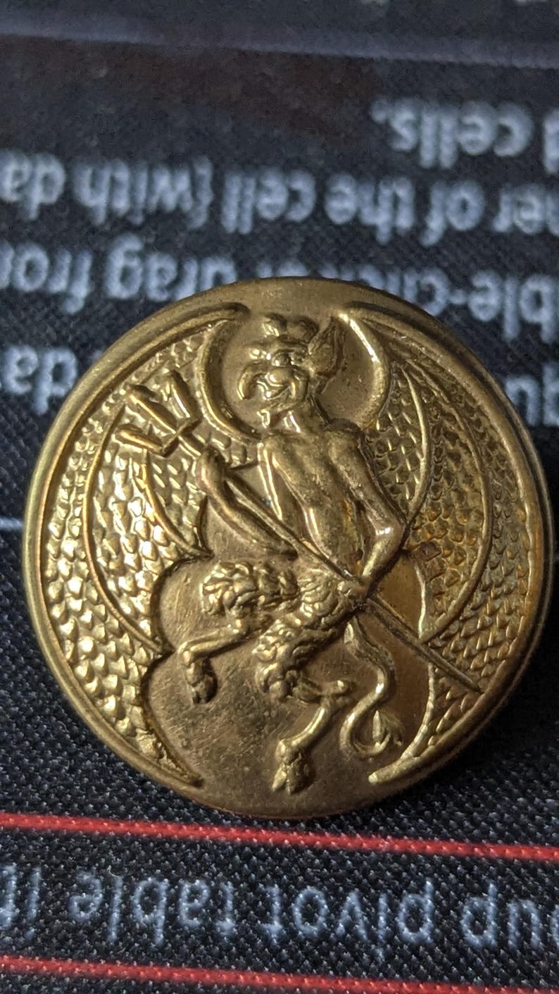 May include: A gold-colored button with a detailed relief of a devil-like figure holding a trident. The button has a circular shape with intricate detailing around the edges and the central image.