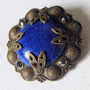 May include: Vintage brooch with a central oval, deep blue stone. The stone is surrounded by an ornate, gold-toned metal frame with decorative leaf accents and small, dark crystals. The brooch has a pin closure on the back.