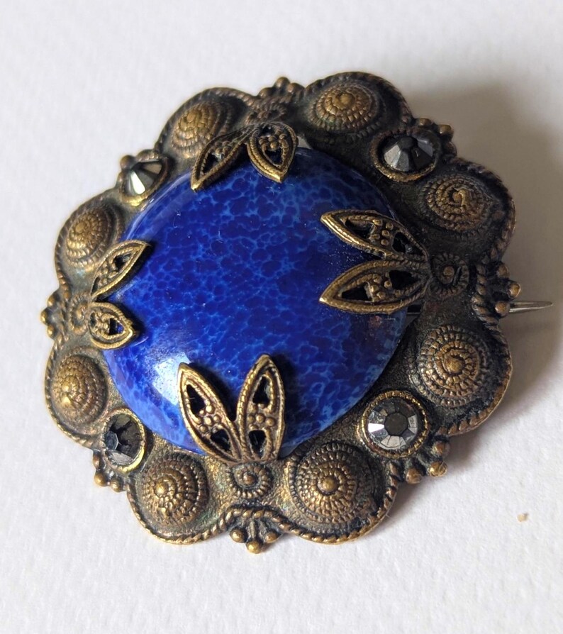 May include: Vintage brooch with a central oval, deep blue stone. The stone is surrounded by an ornate, gold-toned metal frame with decorative leaf accents and small, dark crystals. The brooch has a pin closure on the back.