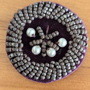 May include: A round, purple velvet brooch with a center design of white beads and a border of small, brown beads.