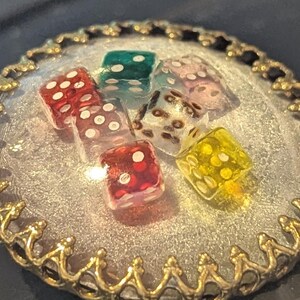May include: A round, silver-colored pendant with a gold-colored, ornate border. The pendant contains six colorful dice with white dots. The dice are red, green, clear, pink, and yellow. The pendant is on a dark surface.