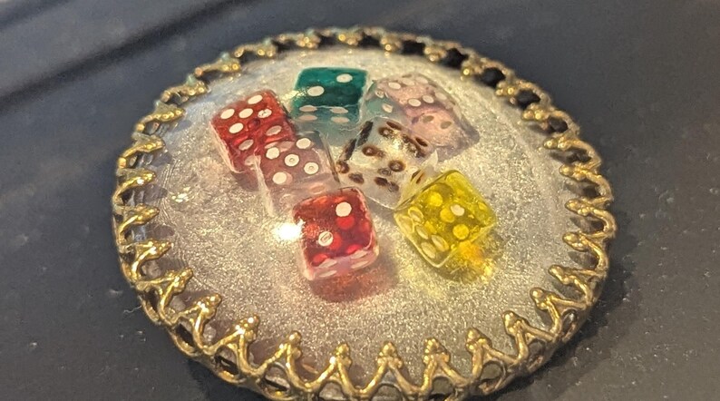 May include: A round, silver-colored pendant with a gold-colored, ornate border. The pendant contains six colorful dice with white dots. The dice are red, green, clear, pink, and yellow. The pendant is on a dark surface.