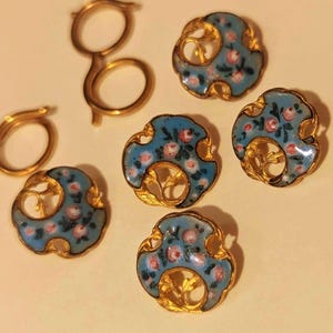 May include: A collection of antique jewelry pieces, including several gold-toned buttons with a light blue enamel floral design and two pairs of gold-toned spectacles. The buttons feature pink and white floral details.