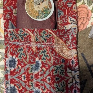 May include: A rectangular fabric pouch featuring a red background with a dense floral and bird pattern in shades of blue, green, and cream. A dark brown wrapped item sits inside, topped with a circular sticker depicting a profile of a person.