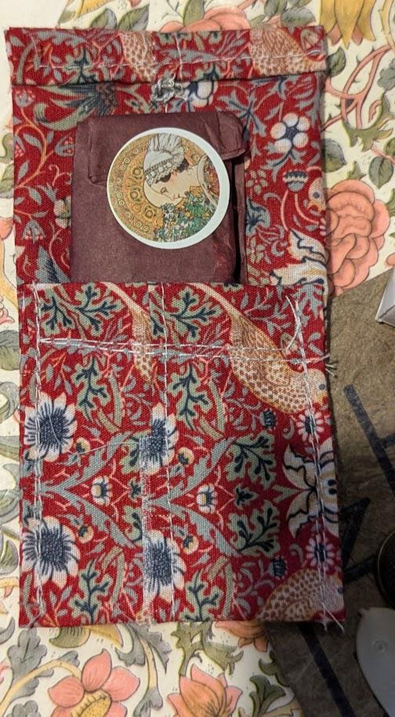May include: A rectangular fabric pouch featuring a red background with a dense floral and bird pattern in shades of blue, green, and cream. A dark brown wrapped item sits inside, topped with a circular sticker depicting a profile of a person.