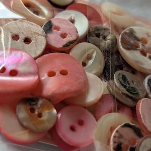 May include: A collection of assorted round buttons in various shades of pink, white, and brown, featuring different textures and finishes. These buttons are two-hole and four-hole styles, packaged together in a clear plastic bag on a pink card.