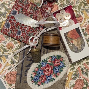 May include: A collection of sewing notions including a red floral fabric pouch, a silver fish-shaped tool, a wooden spool, a brass thimble, a white ring, a box of white buttons, and an oval needlepoint patch featuring a pink and blue floral design.