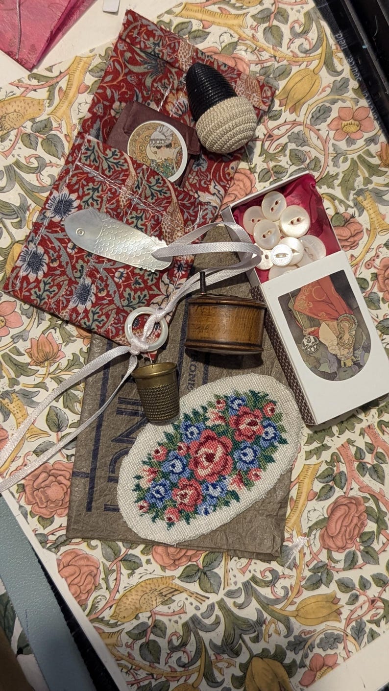 May include: A collection of sewing notions including a red floral fabric pouch, a silver fish-shaped tool, a wooden spool, a brass thimble, a white ring, a box of white buttons, and an oval needlepoint patch featuring a pink and blue floral design.