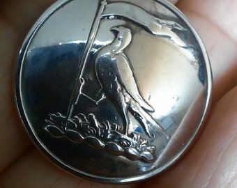 May include: A silver button with a raised design of a bird standing on a rock holding a flag. The bird is facing to the right and the flag is waving in the wind.