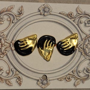Può includere: Tre spille decorative raffiguranti una mano color oro che poggia su una superficie nera lucida. Ogni mano è adornata con un piccolo anello sul dito. Le spille sono esposte su un cartoncino rettangolare decorato con una cornice ovale.