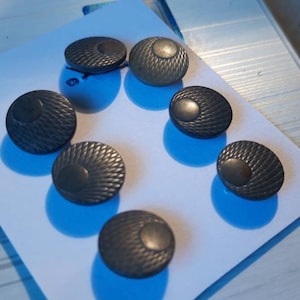 Puede incluir: Una colección de ocho botones de color gris oscuro con un patrón texturizado y tejido. Cada botón tiene un centro elevado y está dispuesto sobre una superficie blanca. Los botones son redondos y parecen ser de metal.