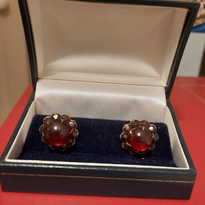 May include: A pair of red gemstone earrings in a blue velvet jewelry box. The earrings are set in gold and feature a round, cabochon-cut gemstone surrounded by smaller gemstones.