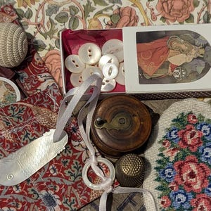 May include: A collection of sewing notions including a white crocheted pincushion, a box of white buttons with a decorative illustration, a wooden thread spool, a metal thimble, a fish-shaped needle threader, and a floral needlepoint patch.