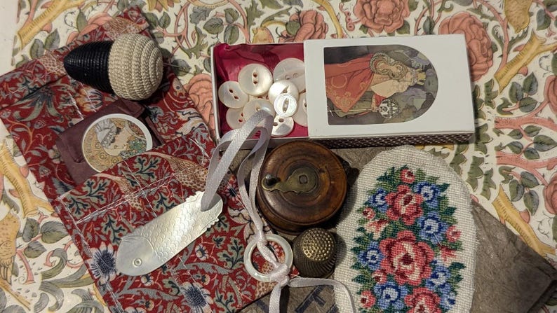 May include: A collection of sewing notions including a white crocheted pincushion, a box of white buttons with a decorative illustration, a wooden thread spool, a metal thimble, a fish-shaped needle threader, and a floral needlepoint patch.