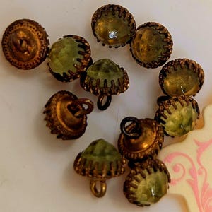 Pode incluir: Uma coleção de botões vintage com uma mistura de estilos. Alguns botões são redondos com acabamento em latão e outros apresentam um centro de vidro verde facetado. Os botões têm uma estética decorativa e antiga.