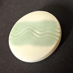 May include: A ceramic, oval-shaped coaster with a two-tone design. The top half features a light green wavy pattern, while the bottom half is a creamy white. The coaster is smooth and appears to be handmade.