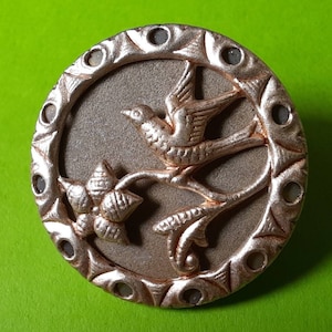 May include: A silver-toned metal button with a raised design of a bird perched on a branch with a flower. The button has a decorative border with a textured finish.