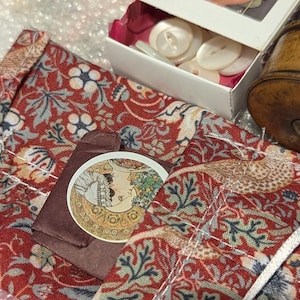May include: A red fabric pouch with a floral pattern featuring blue and green leaves, a circular sticker with an illustration of a person, a white box containing white buttons, and a cylindrical wooden spool with a black thread top.
