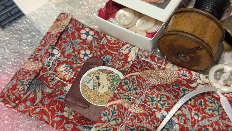 May include: A red fabric pouch with a floral pattern featuring blue and green leaves, a circular sticker with an illustration of a person, a white box containing white buttons, and a cylindrical wooden spool with a black thread top.